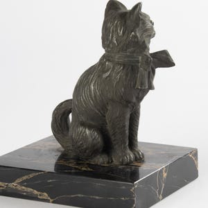 May include: A bronze cat sculpture wearing a scarf and bow, seated on a black marble base with gold veining. The cat has textured fur and is looking to the side. The sculpture is a decorative item.