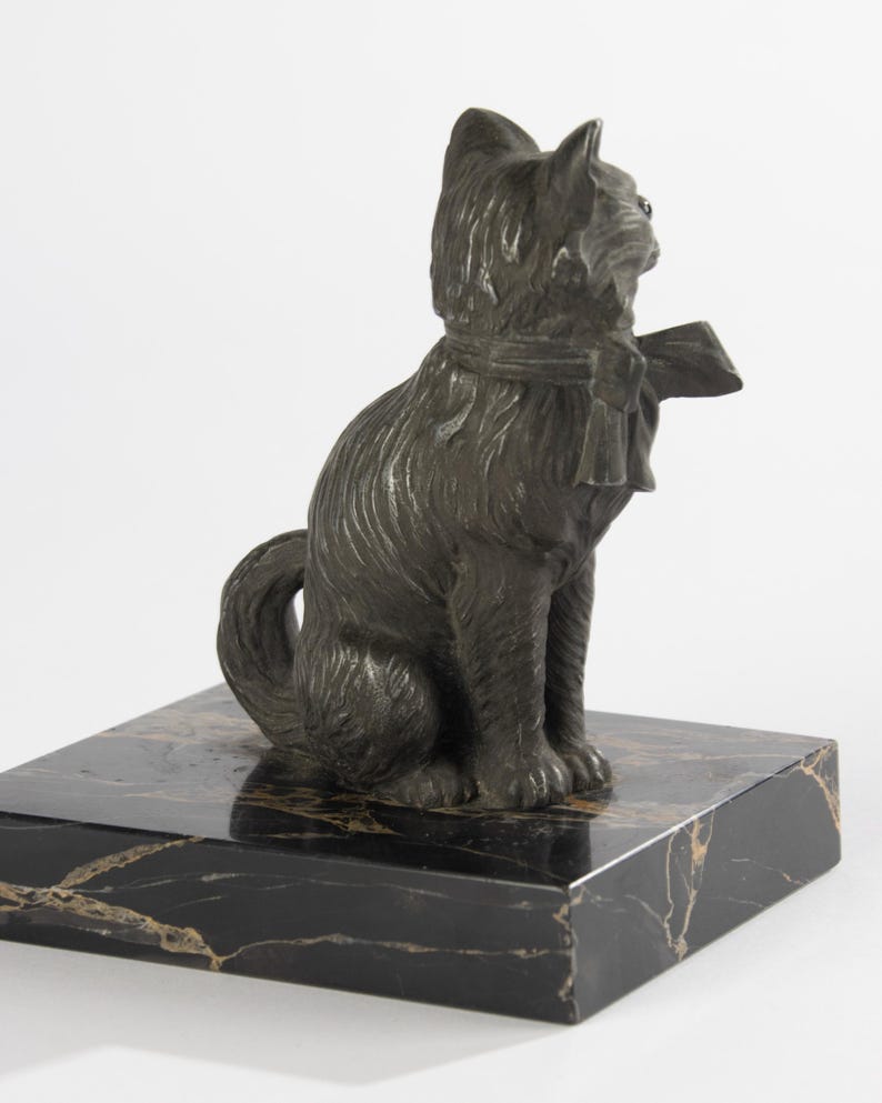 May include: A bronze cat sculpture wearing a scarf and bow, seated on a black marble base with gold veining. The cat has textured fur and is looking to the side. The sculpture is a decorative item.