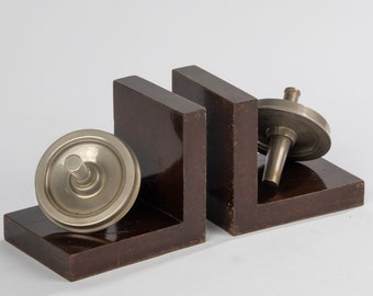 Puede incluir: Un par de sujetalibros de madera oscura con tapas de metal plateado en forma de peonzas. Los sujetalibros tienen forma de L y están sobre una superficie blanca.
