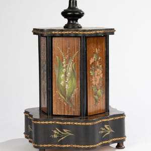 May include: A hexagonal wooden music box featuring a black finish with gold-painted trim and a central finial. The side panels display hand-painted floral motifs, including lily of the valley and pink blossoms, set against a wood-grain background.