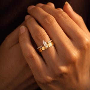Könnte beinhalten: Ein Goldring-Set bestehend aus einem breiten, polierten Goldring und einem schmaleren, mit Diamanten im Pavé-Stil besetzten Ring. Der breitere Ring hat einen zentralen Solitärdiamanten im Marquise-Schliff, was einen geschichteten Look erzeugt.