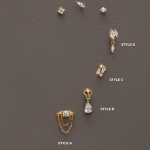 Könnte beinhalten: Eine Auswahl an goldfarbenen Ohrringen mit verschiedenen Designs, darunter Ohrstecker und Hängeohrringe. Die Ohrringe verfügen über klare Edelsteine in verschiedenen Formen, wie rund und tropfenförmig. Die Ohrringe werden auf einer grauen Oberfläche präsentiert.