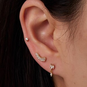 Könnte beinhalten: Nahaufnahme eines Ohrs mit drei goldenen Ohrringen. Die Ohrringe bestehen alle aus kleinen, klaren Steinen. Der obere Ohrring ist ein einfacher Stecker, der mittlere Ohrring ist eine gebogene Stange mit Steinen und der untere Ohrring ist ein Hängeohrring mit einer Gruppe von Steinen.