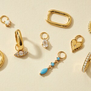 Könnte beinhalten: Eine Sammlung goldfarbener Schmuckanhänger mit verschiedenen Designs, darunter ein Tropfen, ein Herz, ein Auge und ein türkisfarbener Stein. Die Anhänger sind so konzipiert, dass sie an Ohrringe, Halsketten oder Armbänder hinzugefügt werden können.