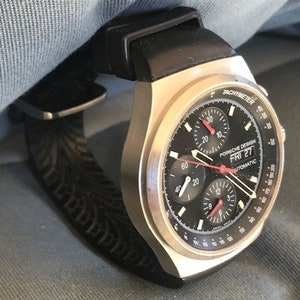 May include: A silver-toned Porsche Design watch with a black face and a black rubber strap. The watch face displays multiple sub-dials, red hands, and the text "PORSCHE DESIGN FRI 27 AUTOMATIC". The watch is on a denim background.