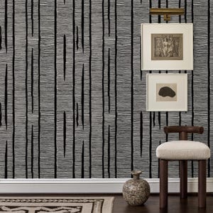 Può includere: Un interno con carta da parati a righe bianche e nere, due stampe d'arte incorniciate appese alla parete, una luce per quadri in ottone, un vaso in ceramica rotondo e testurizzato e uno sgabello in legno con seduta imbottita color crema.