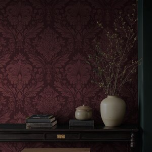 May include: A burgundy damask wallpaper forms the backdrop for a black table holding a stack of books, a small ceramic jar with a lid, and a cream-colored vase with dried branches. The scene evokes a sense of classic elegance.