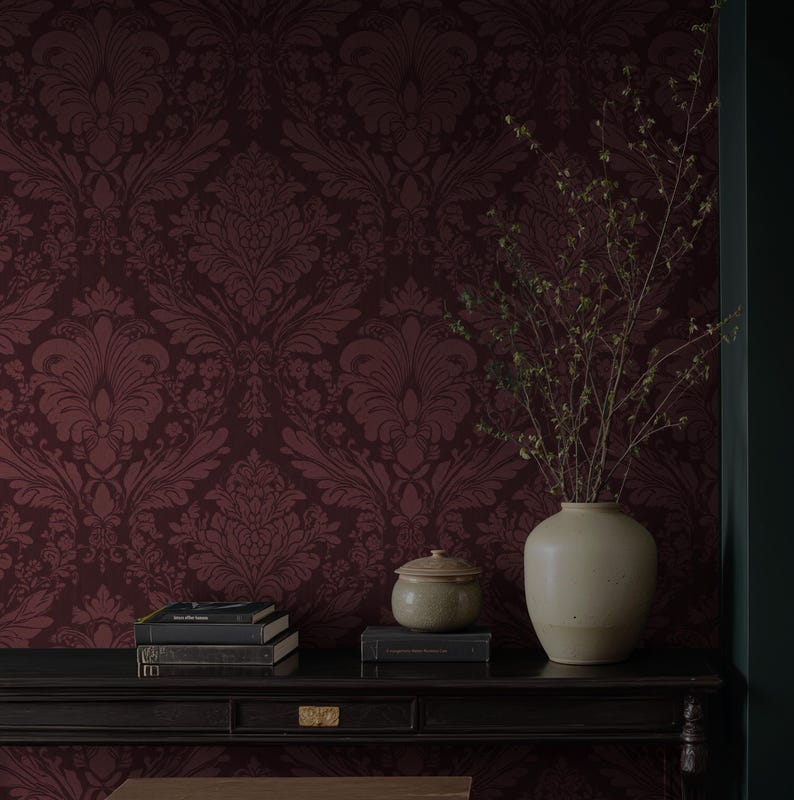 May include: A burgundy damask wallpaper forms the backdrop for a black table holding a stack of books, a small ceramic jar with a lid, and a cream-colored vase with dried branches. The scene evokes a sense of classic elegance.
