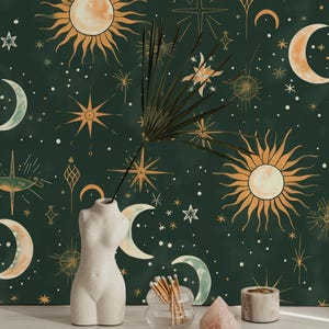 Op de afbeelding: Een hemels thema met een donkergroene achtergrond versierd met gouden en crèmekleurige zonnen, manen en sterren. Een witte, torso-vormige vaas houdt een bladstengel vast. Andere elementen zijn een glazen pot met lucifers, een roze kristal en een kleine kaarsenhouder.