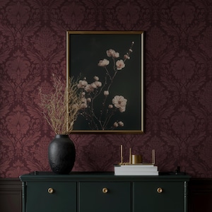 May include: A dark green cabinet with gold knobs and a black vase holding dried branches. A framed floral print with a gold frame hangs on a burgundy damask wallpaper background. A gold candle holder and books are on the cabinet.