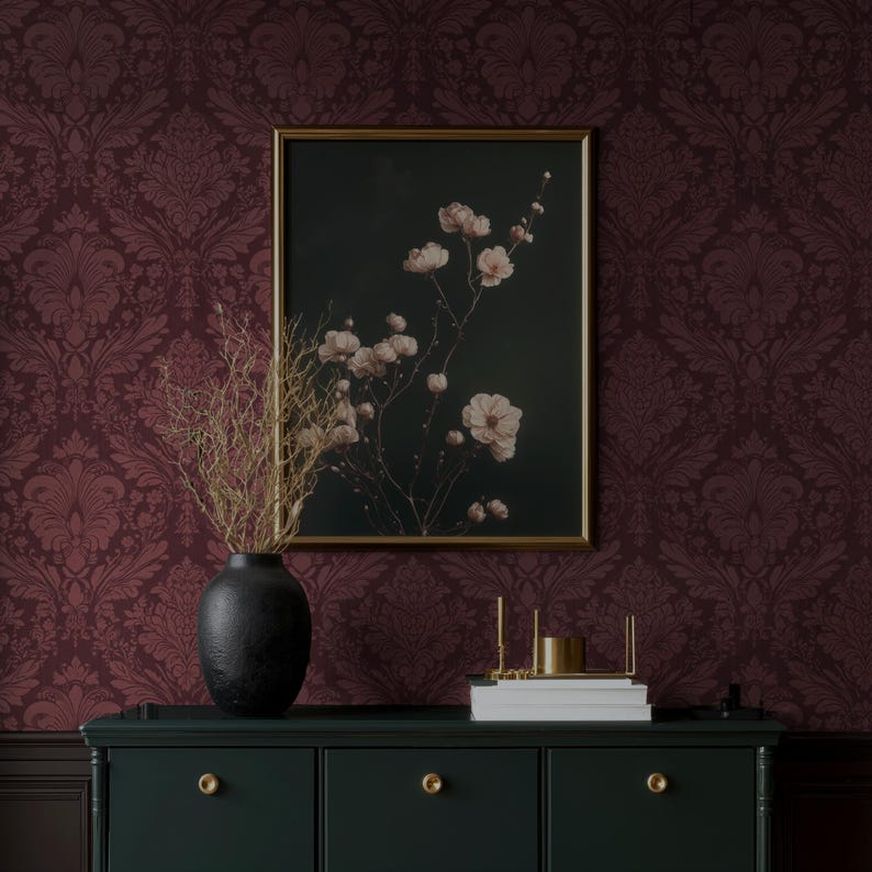 May include: A dark green cabinet with gold knobs and a black vase holding dried branches. A framed floral print with a gold frame hangs on a burgundy damask wallpaper background. A gold candle holder and books are on the cabinet.