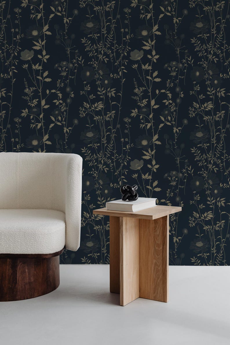 May include: Dark navy wallpaper with a botanical design featuring beige-colored flowers and foliage. A white armchair with a wooden base and a small wooden side table with a book and decorative object are in the foreground.