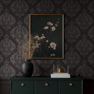 May include: A framed floral print with a gold frame hangs above a dark green cabinet. A black vase holds dried branches. The cabinet has gold knobs and a stack of books with a gold candle holder.