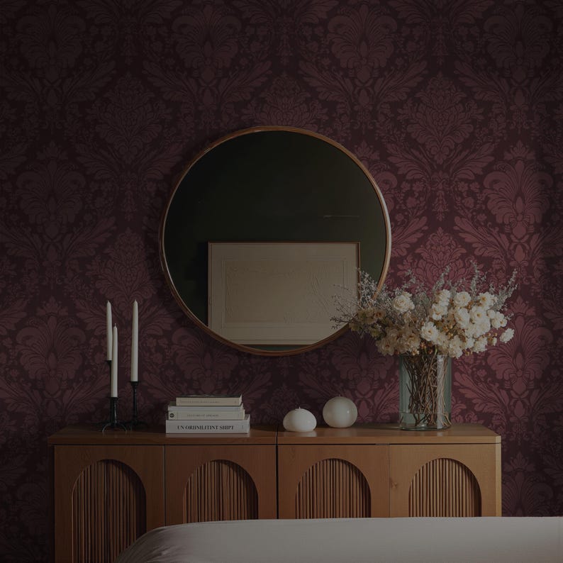 May include: A room with a burgundy damask wallpaper design. A round mirror hangs above a wooden cabinet with books, candles, and a vase of flowers. The room has a warm, inviting atmosphere.