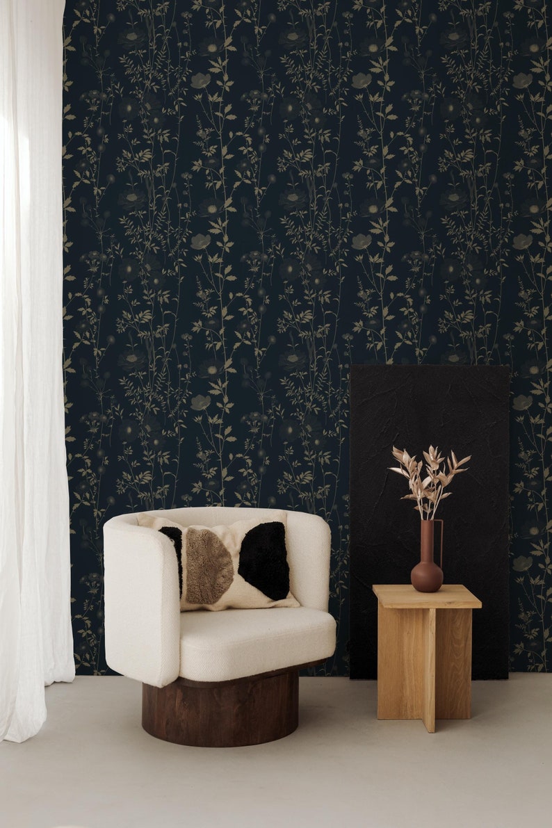 May include: A cozy interior scene featuring a white armchair with a dark wood base, a patterned pillow, and a small wooden side table. The backdrop is a dark blue floral wallpaper. A vase with dried flowers sits on the table.