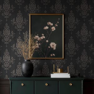 May include: A dark green dresser with gold hardware supports a black vase with dried branches and a framed floral print. The background features a dark wallpaper with a repeating pattern.