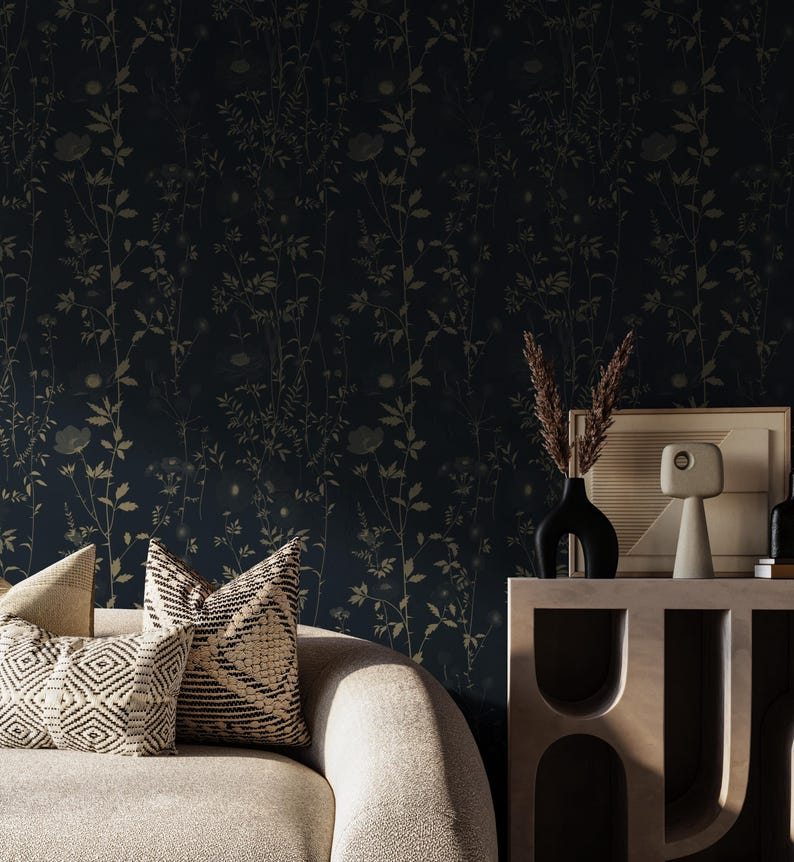 May include: A dark blue floral wallpaper with a repeating pattern of gold-colored flowers and foliage. Two decorative pillows with geometric patterns rest on a light-colored sofa. A black vase and a framed picture sit on a wooden table.