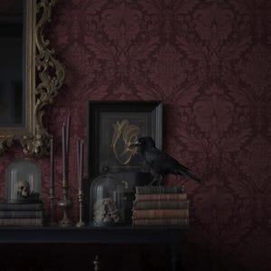 May include: A dark, gothic-themed display featuring a black raven perched on antique books. The scene includes a skull in a glass dome, candles, and a framed artwork, all set against a burgundy damask wallpaper. A decorative mirror is on the left.