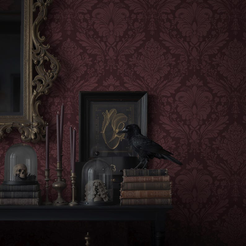 May include: A dark, gothic-themed display featuring a black raven perched on antique books. The scene includes a skull in a glass dome, candles, and a framed artwork, all set against a burgundy damask wallpaper. A decorative mirror is on the left.