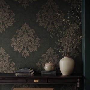 May include: A dark green wall with a repeating brown damask pattern. A dark wooden table holds a stack of books, a small ceramic jar with a lid, and a cream-colored vase with branches.