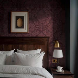 May include: A bedroom scene featuring a dark wood headboard, white pillows, and a striped duvet. A framed artwork with a gold egg is mounted on a patterned burgundy wall. A bedside table holds a lamp and a cup of coffee.