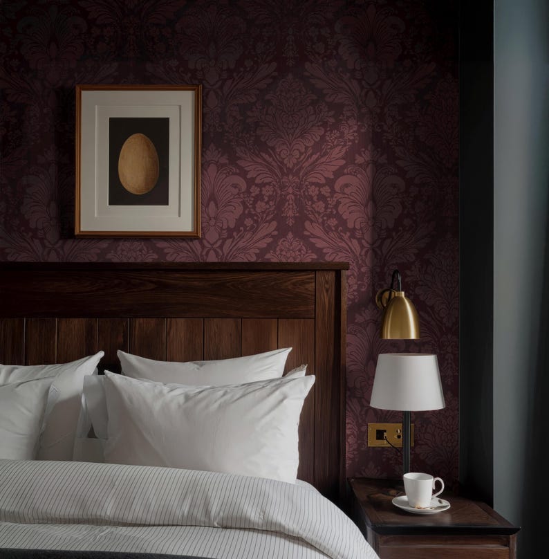 May include: A bedroom scene featuring a dark wood headboard, white pillows, and a striped duvet. A framed artwork with a gold egg is mounted on a patterned burgundy wall. A bedside table holds a lamp and a cup of coffee.