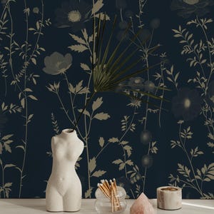 May include: A still life arrangement featuring a white, torso-shaped vase, a glass container with matches, a pink crystal, and a small candle holder. The background is a dark blue floral wallpaper with beige botanical designs.