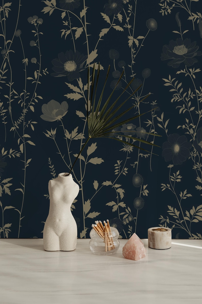 May include: A still life arrangement featuring a white, torso-shaped vase, a glass container with matches, a pink crystal, and a small candle holder. The background is a dark blue floral wallpaper with beige botanical designs.