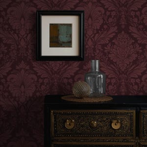 May include: A framed abstract painting hangs on a burgundy damask wallpapered wall. A black cabinet with gold accents holds a clear glass bottle, a decorative sphere, and a woven mat.