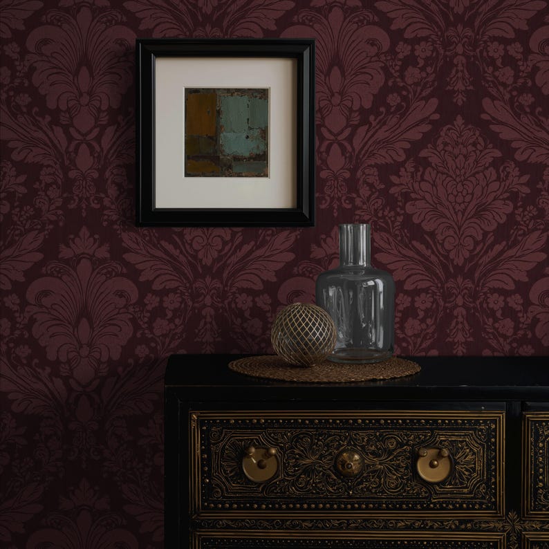 May include: A framed abstract painting hangs on a burgundy damask wallpapered wall. A black cabinet with gold accents holds a clear glass bottle, a decorative sphere, and a woven mat.