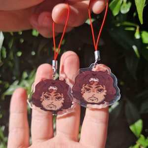 Op de afbeelding: Twee heldere acryl bedels met een cartoon illustratie van een persoon met bruin haar en een baard. Elke bedel heeft een rood touwtje en een zilverkleurige metalen sluiting. De illustratie heeft een rode edelsteen op het voorhoofd.