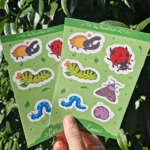 Op de afbeelding: Twee groene sticker vellen met kleurrijke insecten illustraties, waaronder een lieveheersbeestje, rups en kever. De stickers hebben een witte rand en zijn geplaatst tegen een achtergrond van groene bladeren. Tekst op de stickers luidt "Critter Crew" en "by DoodlDoot".