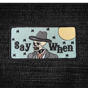 May include: Embroidered patch featuring a skeleton wearing a hat and suit, with the words "Say When." The design includes a yellow moon and small black crosses on a light blue background.