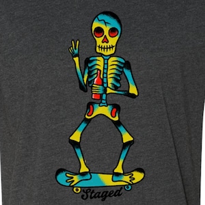 May include: A dark heather gray t-shirt featuring a graphic of a yellow and blue skeleton riding a skateboard. The skeleton holds a red bottle in one hand and makes a peace sign with the other. The word Staged is printed in black below the skateboard.