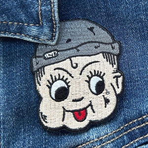 May include: A beige embroidered patch of a cartoon character with a gray hat, big eyes, and a red tongue sticking out. The patch is sewn onto a blue denim jacket.