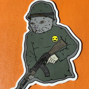 May include: A sticker of a cat wearing a military helmet and holding a rifle. The cat is smoking a cigarette and has a smiley face sticker on its uniform.