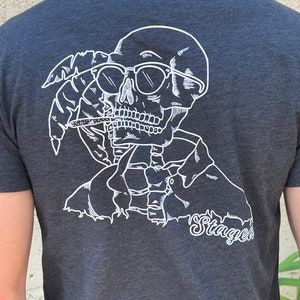 May include: A dark gray t-shirt with a white graphic of a skull wearing sunglasses and holding a cigarette. The skull is surrounded by palm leaves. The text "Staged" is printed below the skull.