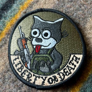 May include: Embroidered patch featuring a cartoon cat with a rifle, wearing a military vest, and sticking out its tongue. The patch is circular with the words "LIBERTY OR DEATH" in a circular pattern around the cat.
