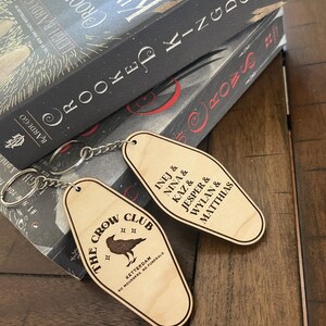 Peut inclure: Deux porte-cl&eacute;s en bois en forme de cl&eacute;s de chambre d'h&ocirc;tel. Un porte-cl&eacute;s pr&eacute;sente le texte "The Crow Club" avec une illustration de corbeau et le texte "Ketterdam, No Mourners, No Funerals". L'autre porte-cl&eacute;s pr&eacute;sente le texte "Inej & Nina & Kaz & Jesper & Wylan & Matthias".
