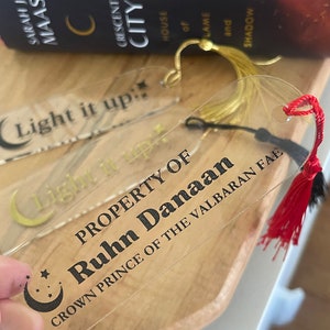 Op de afbeelding: Een transparante acryl boekenlegger met een gouden kwast en een zwart halve maan ontwerp. De boekenlegger heeft de tekst "Light it up" en "PROPERTY OF Ruhn Danaan CROWN PRINCE OF THE VALBARAN FAE" erop gedrukt.