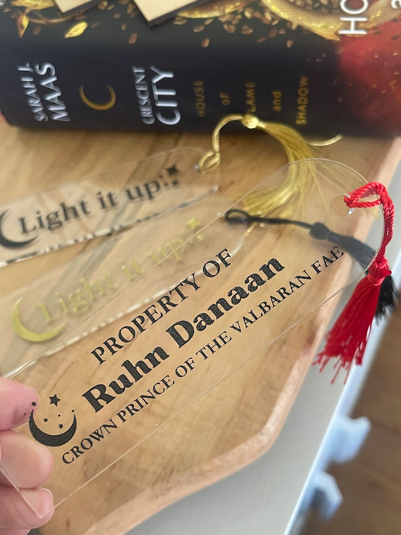 Op de afbeelding: Een transparante acryl boekenlegger met een gouden kwast en een zwart halve maan ontwerp. De boekenlegger heeft de tekst "Light it up" en "PROPERTY OF Ruhn Danaan CROWN PRINCE OF THE VALBARAN FAE" erop gedrukt.