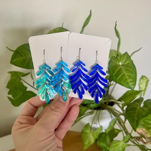 May include: Earrings with a leaf design in shades of blue and teal. The earrings are displayed on white cards with silver hooks. The leaves have a glossy finish and are arranged in a layered pattern. The earrings are a unique accessory.