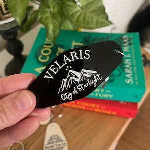May include: Black key chain with a white design of mountains and stars. The text "Velaris City of Starlight" is engraved on the keychain.