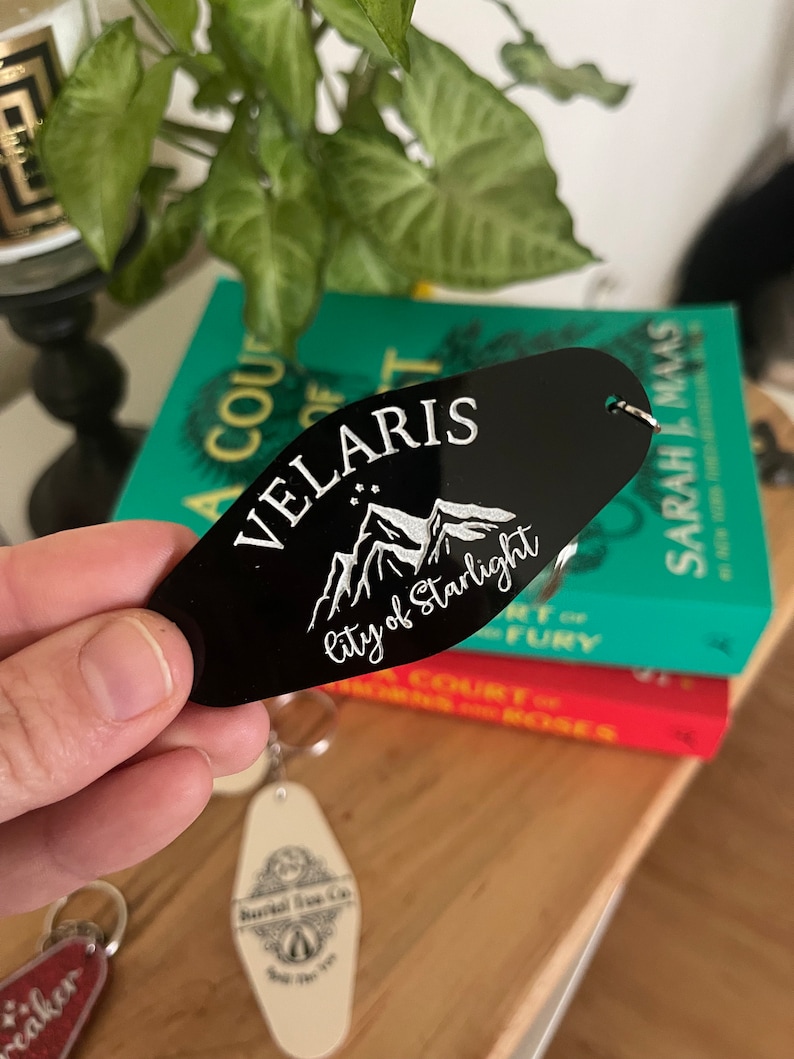 May include: Black key chain with a white design of mountains and stars. The text "Velaris City of Starlight" is engraved on the keychain.