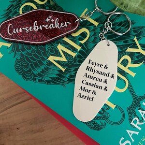 May include: A white keychain with the text "Feyre & Rhysand & Amren & Cassian & Mor & Azriel" on it. The keychain is attached to a silver key ring and is sitting on top of a green book with the text "A Court of Mist and Fury" on the cover.