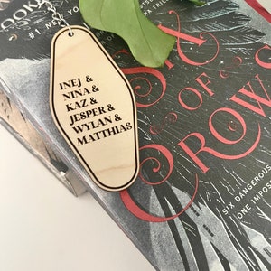 Peut inclure: Porte-cl&eacute;s en bois en forme de cl&eacute; d'h&ocirc;tel avec le texte "INEJ & NINA & KAZ & JESPER & WYLAN & MATTHIAS" dessus. Le porte-cl&eacute;s est pos&eacute; sur un livre dont la couverture porte le titre "Six of Crows".