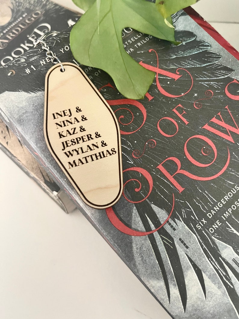 Peut inclure: Porte-cl&eacute;s en bois en forme de cl&eacute; d'h&ocirc;tel avec le texte "INEJ & NINA & KAZ & JESPER & WYLAN & MATTHIAS" dessus. Le porte-cl&eacute;s est pos&eacute; sur un livre dont la couverture porte le titre "Six of Crows".