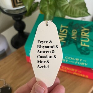 May include: White motel key tag with black text listing the names Feyre, Rhysand, Amren, Cassian, Mor, and Azriel. The key tag is held in a hand with a book titled "A Court of Mist and Fury" in the background.
