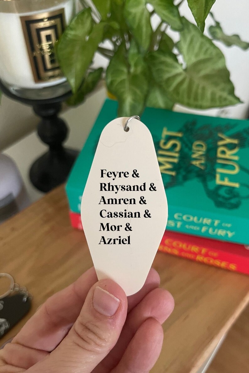 May include: White motel key tag with black text listing the names Feyre, Rhysand, Amren, Cassian, Mor, and Azriel. The key tag is held in a hand with a book titled "A Court of Mist and Fury" in the background.