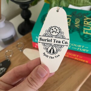 May include: White motel key tag with black text and a logo. The text reads "Suriel Tea Co. Spill The Tea".